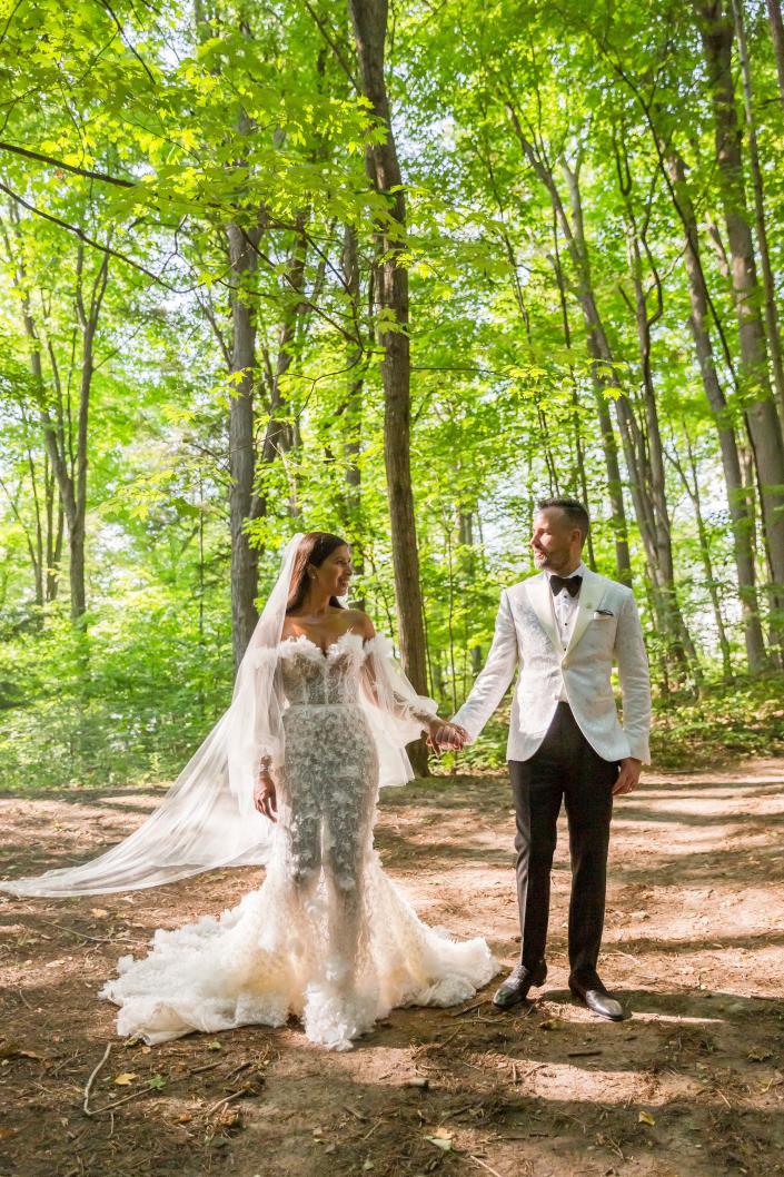 A bride and groom look at each other and hold hands in their wedding attire in a forest.