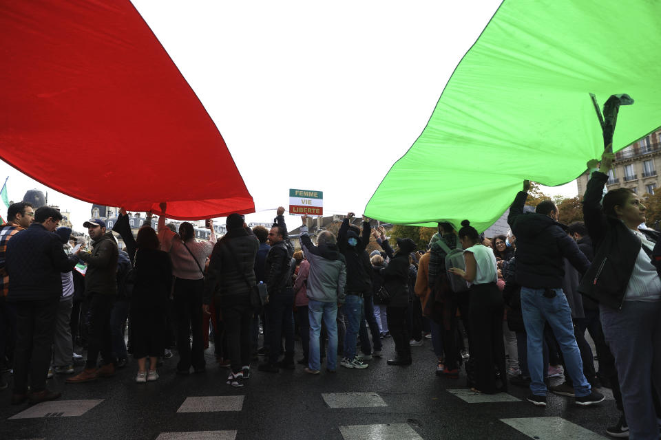 Protesters stand under an Iranian flag during a demonstration to show support for Iranian protesters standing up to their leadership over the death of a young woman in police custody, Sunday, Oct. 2, 2022 in Paris. Thousands of Iranians have taken to the streets over the last two weeks to protest the death of Mahsa Amini, a 22-year-old woman who had been detained by Iran's morality police in the capital of Tehran for allegedly not adhering to Iran's strict Islamic dress code. (AP Photo/Aurelien Morissard)