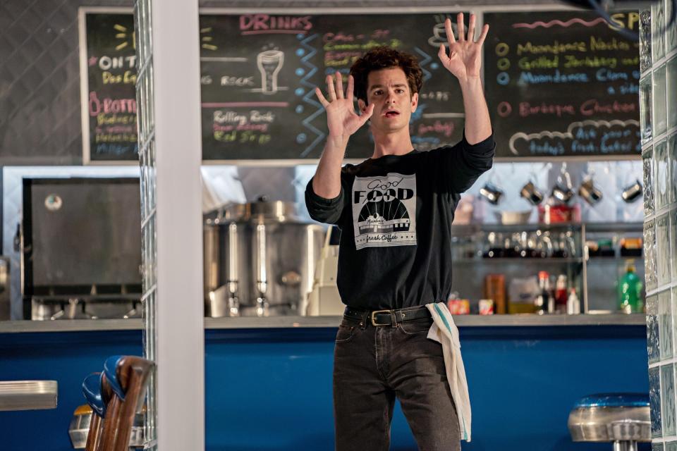 A white man with frizzy brown hair stands with his back to a diner counter. He looks outside in amazement. His hands are raised.