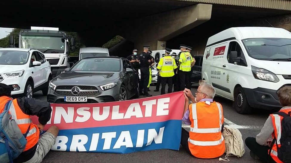 Protesters hold banner in from of vans and cars