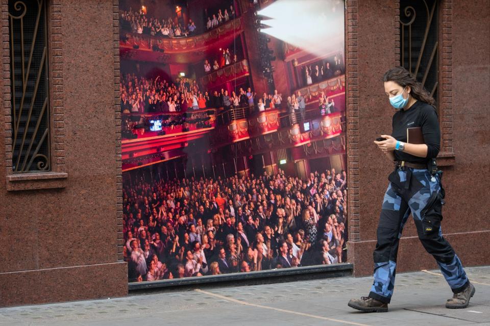 <p>A woman wearing a face mask passes a photo of a packed theatre auditorium in Soho</p> (PA)