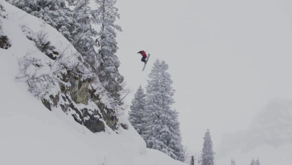 A skier leaping off a snowy rock face, in midair, two dozen feet above the snow beneath him.