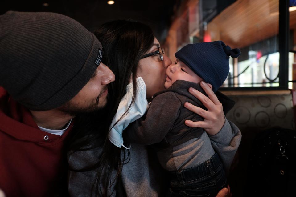 A woman holds a baby, kissing it on the cheek, as a man looks on.