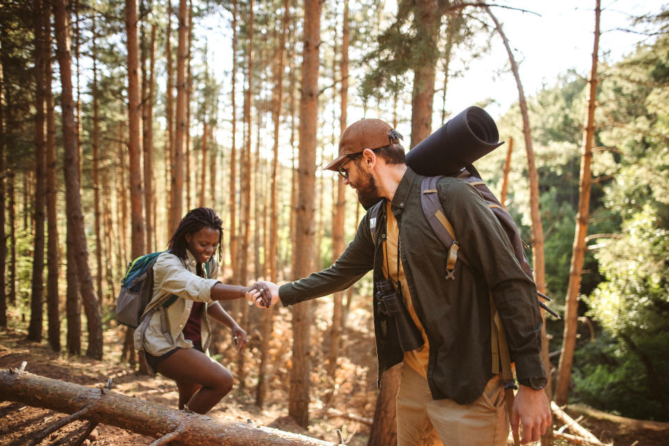 A man and a woman wearing hiking gear help each other while crossing a log in a forest. They both have backpacks and appear to be enjoying an outdoor adventure