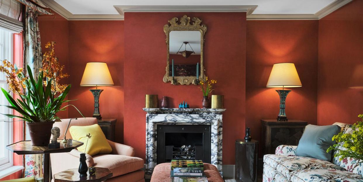 the deep red drawing room has a light gray carpet, two upholstered chairs with accent tables by a window, a marble fireplace with a mirror above it, a large fabric covered ottoman, and a floral sofa
