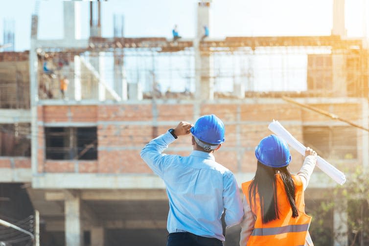 A man and woman wear hardhats looking at the construction of a building.