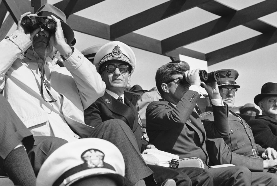 FILE - In this April 14, 1962 file photo, the Shah of Iran, Mohammad Reza Pahlavi sits between President John Kennedy and Vice President Lyndon Johnson as they look through binoculars of Marine landing operations at Camp LeJeuene, North Carolina. The fall of the Peacock Throne and the rise of the Islamic Revolution in Iran grew out of the shah’s ever-tightening control over the country as other Middle East monarchies toppled. (AP Photo, File)