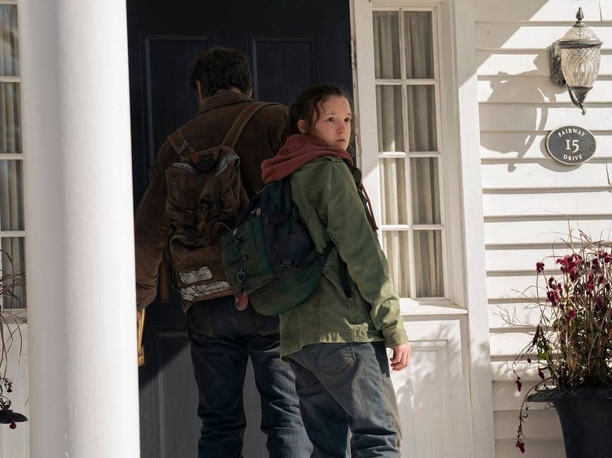 pedro pascal and bella ramsey as joel and ellie in the last of us, entering bill's house in episode three. joel is a middle aged man wearing a brown backpack and jacket, and ellie is a teenager wearing a green backpack and jacket. they're entering a pristine white house with wilted flowers out front