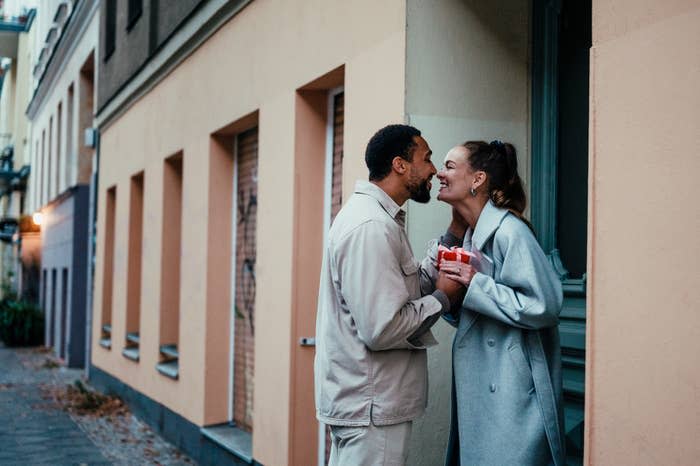 Two people holding drinks stand close, touching foreheads and smiling on a city sidewalk