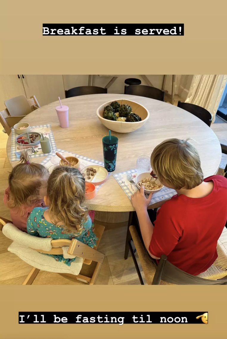 the kid's have their backs to the camera as they sit at the kitchen table with breakfast in front of them