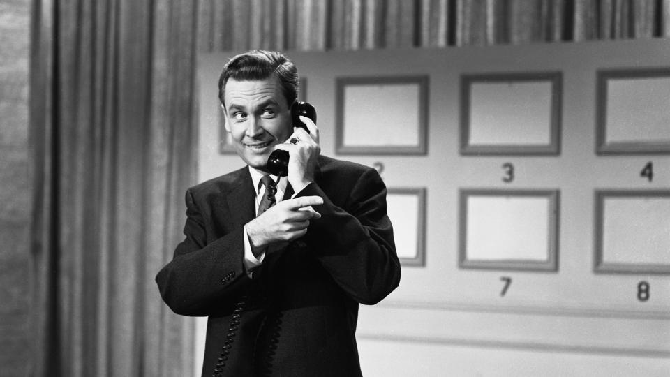 a black and white photo of bob barker on the set of truth or consequences, listening into a telephone and pointing off camera