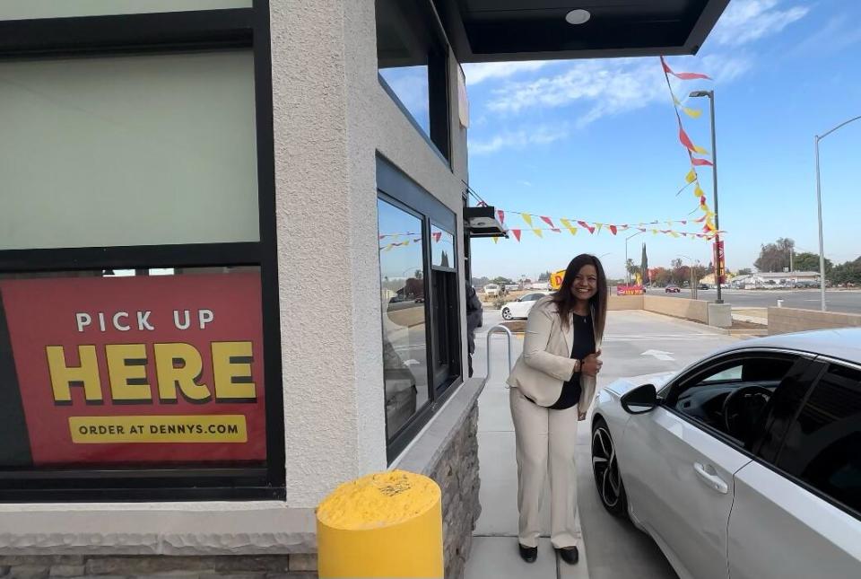 A woman in a white jacket and pants smiles while standing outside a drive-through window, next to a white car