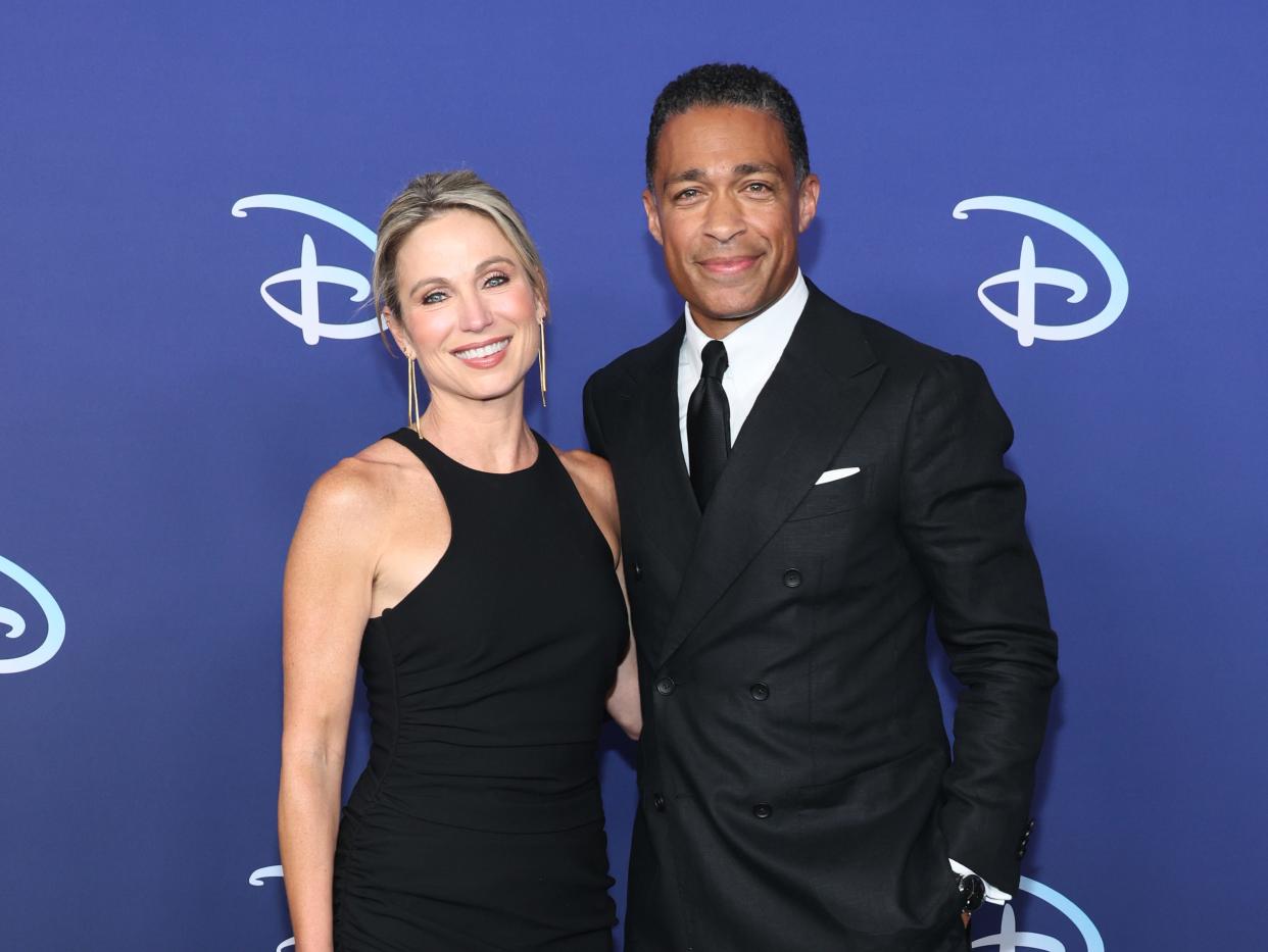 amy robach, wearing a blach dress with an asymmetric, ruffled hem, and t.j. holmes, wearing a black suit and black tie, standing together in front of a navy background with the Disney logo