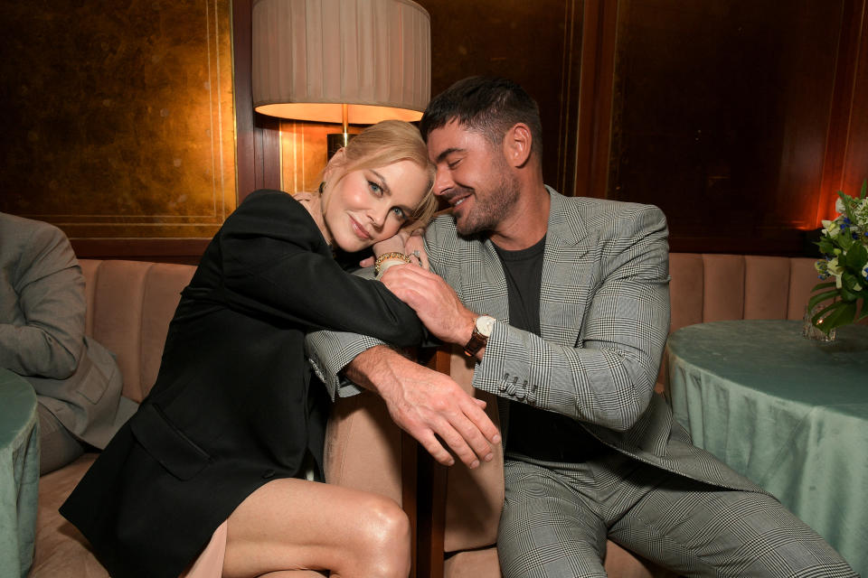 Nicole Kidman and Zac Efron are sitting closely together on a sofa. Kidman, in a blazer and dress, leans on Efron, who is wearing a suit. Both are smiling