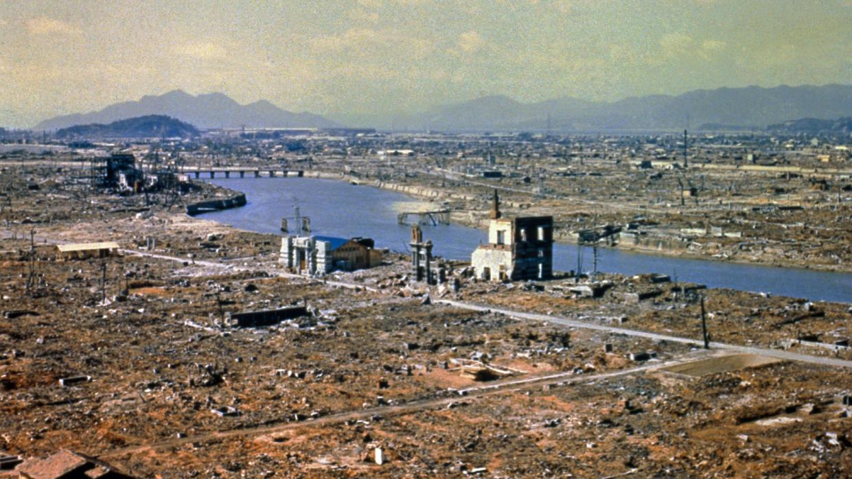 The destruction caused by the nuclear explosion over Hiroshima. The landscape is largely flattened with visible debris from properties. The shells of some properties near a wide river as visible. The ground looks brown.