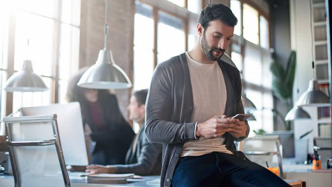 Businessman checking the mobile phone sitting on his desk, colleagues working in the background.