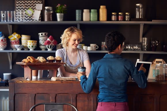 A woman in a bakery smiles from behind the counter as a customer completes her purchase on a point-of-sale terminal.