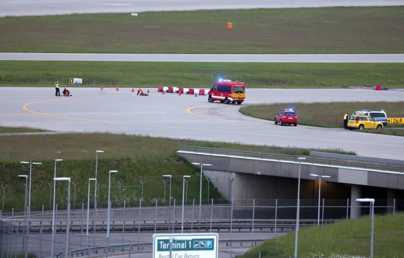 Police and firefighters stand on a runway access road at Franz-Josef-Strauss Airport around climate activists who have stuck themselves there. Climate protection activists paralysed Munich Airport early 18 May. The activists had reached the inner area of the airport grounds. According to their own statements, members of the 'Last Generation' activism group had planned to enter the airport grounds in order to block at least one of the two runways. Karl-Josef Hildenbrand/dpa