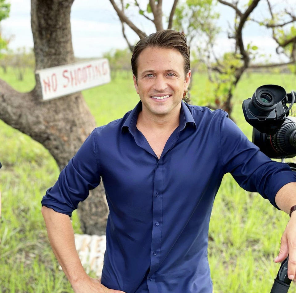 Weekend Sunrise presenter Matt Doran with a TV camera in the countryside. He wears a deep blue shirt with sleeves rolled up.