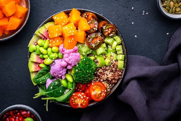 A colorful salad bowl with a variety of vegetables and grains, including butternut squash, tomatoes, edamame, purple cauliflower, quinoa, and broccoli