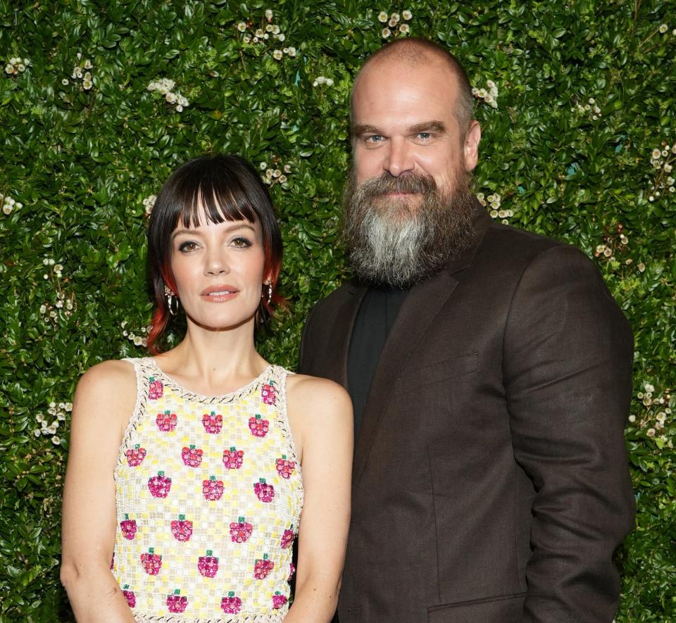 Lily Allen in a floral sleeveless top and striped skirt stands next to David Harbour in a dark suit, posing in front of a greenery backdrop