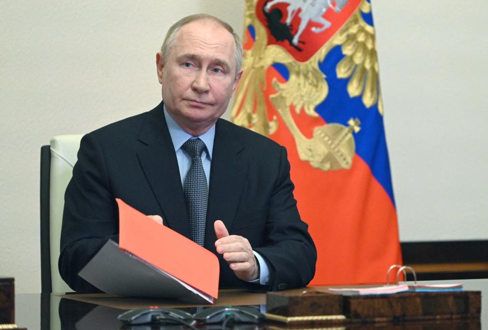 Russian President Vladimir Putin holding paperwork while sitting in a chair in front of the Russian flag.