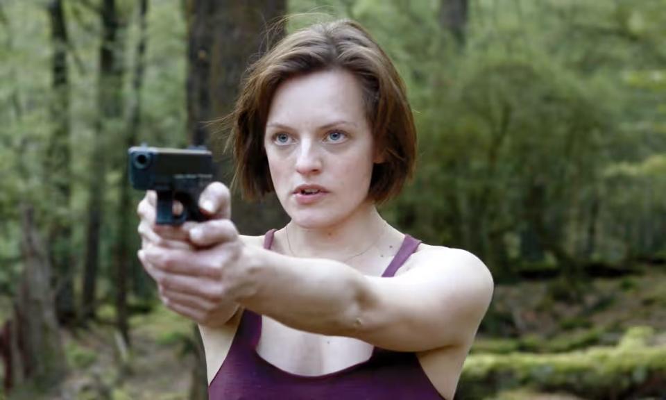 Elisabeth Moss stands straight ahead holding gun.