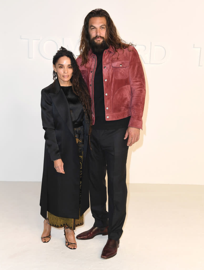 Jason Momoa in a jacket and pants stands next to Lisa Bonet in a long coat, top, and patterned skirt