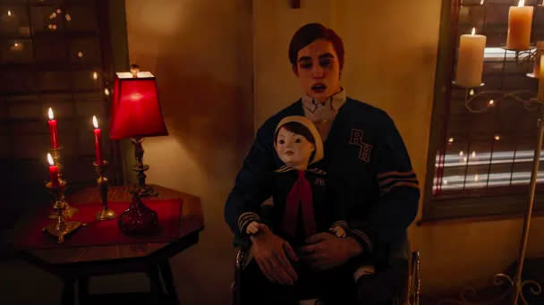 Jason's dead body sits in a wheelchair. He has red hair, his eyes are closed and his mouth is open. He is wearing a shirt and cardigan. He is holding a creepy doll. The room is lit with candles.