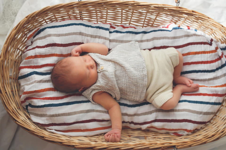 A newborn baby sleeps peacefully in a wicker bassinet lined with a striped blanket. The baby is wearing a light knit top and matching shorts