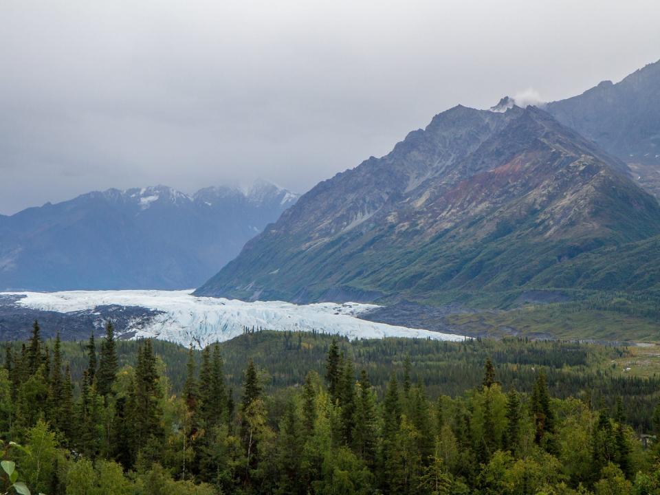 Mountains and a glacier under a cloudy sky surrounded by green trees.