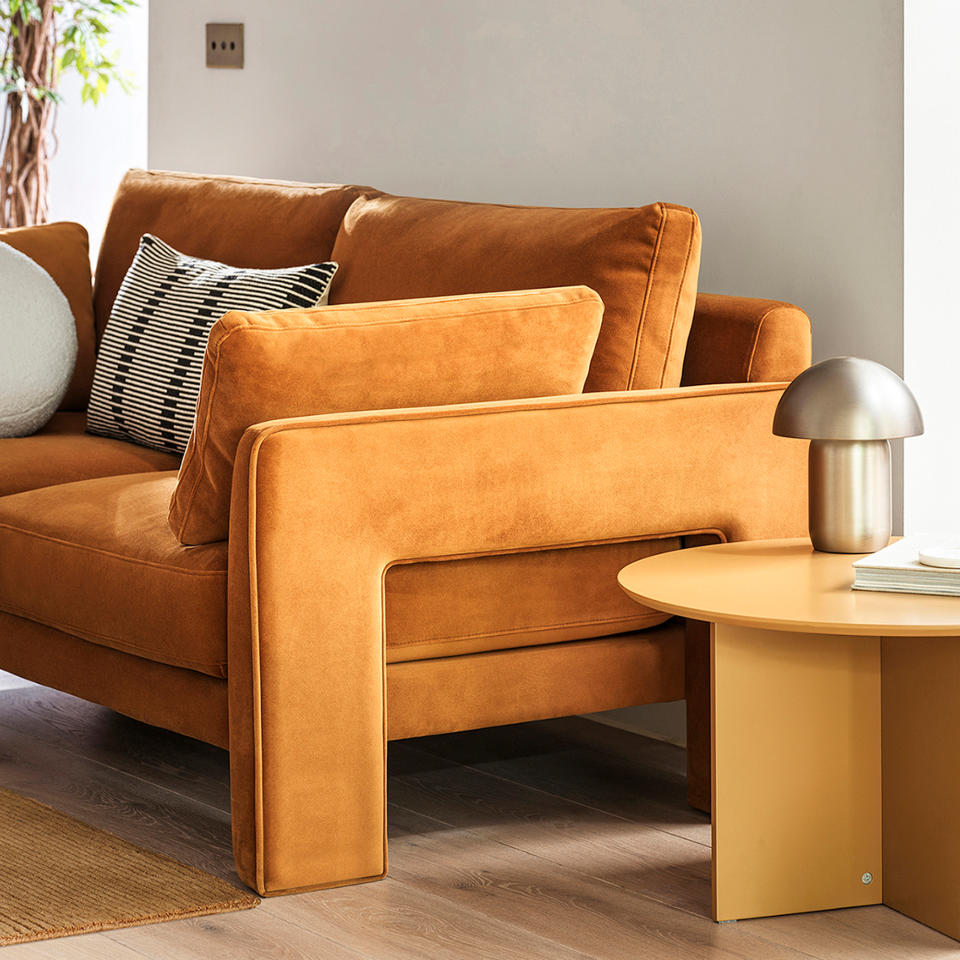 Caramel coloured velvet sofa with blocky style arms on pale wood floor against a white wall