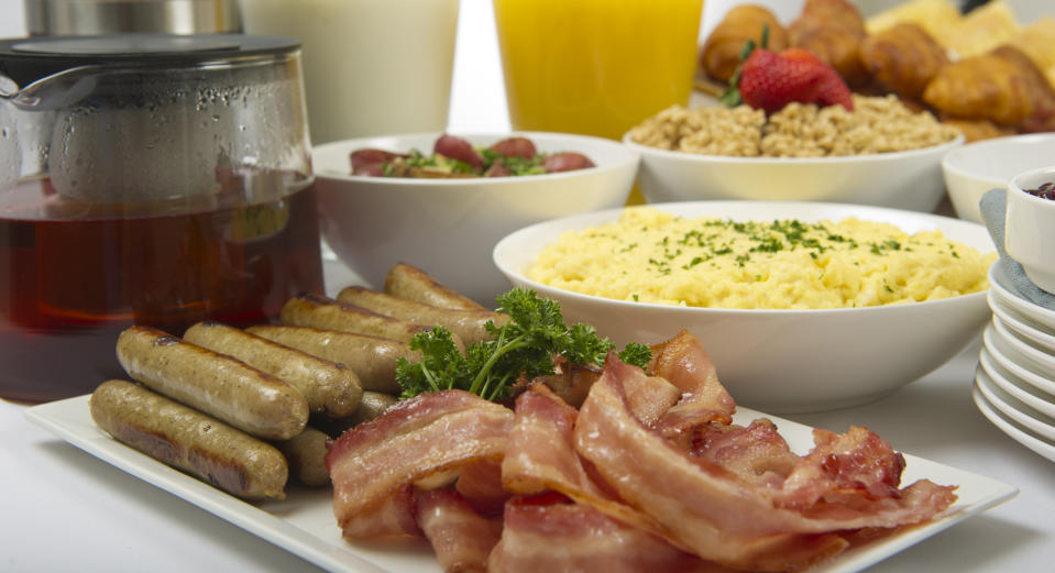 A hearty breakfast spread featuring bacon, sausages, scrambled eggs, croissants, fresh fruit, juice, and a pot of tea