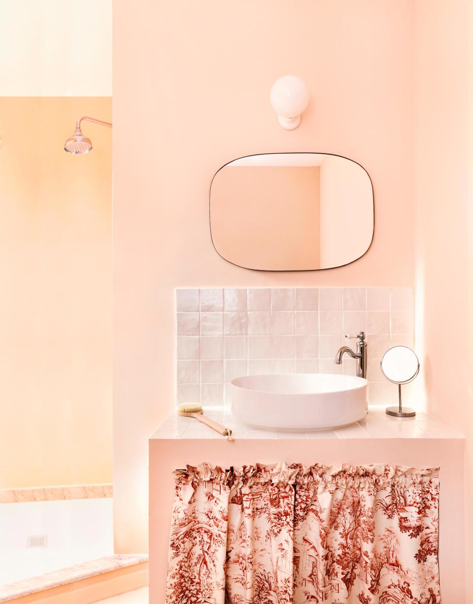 Mirta reimagined the primary bathroom as a relaxing dreamscape with soft pink walls, a lunar sink, Portuguese marble flooring, and a pristine backsplash. The curtain under the sink is a hat-tip to the French countryside. The mirror is from Zara Home.