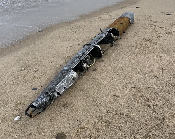 A cylindrical piece of metal looks muddy and rusted as it lies on the sand near the water on a beach.