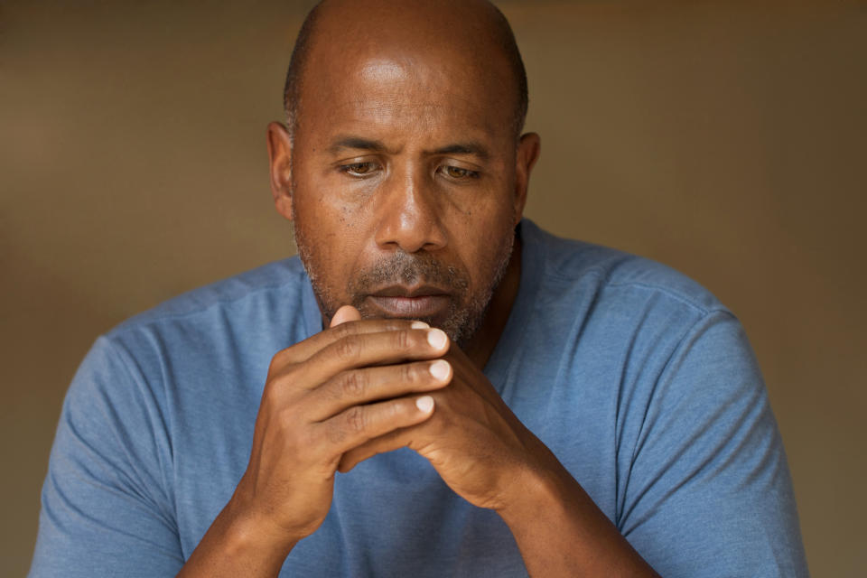 A sullen-looking man, with his hands clasped in front of him, wearing a blue shirt against a brown background.