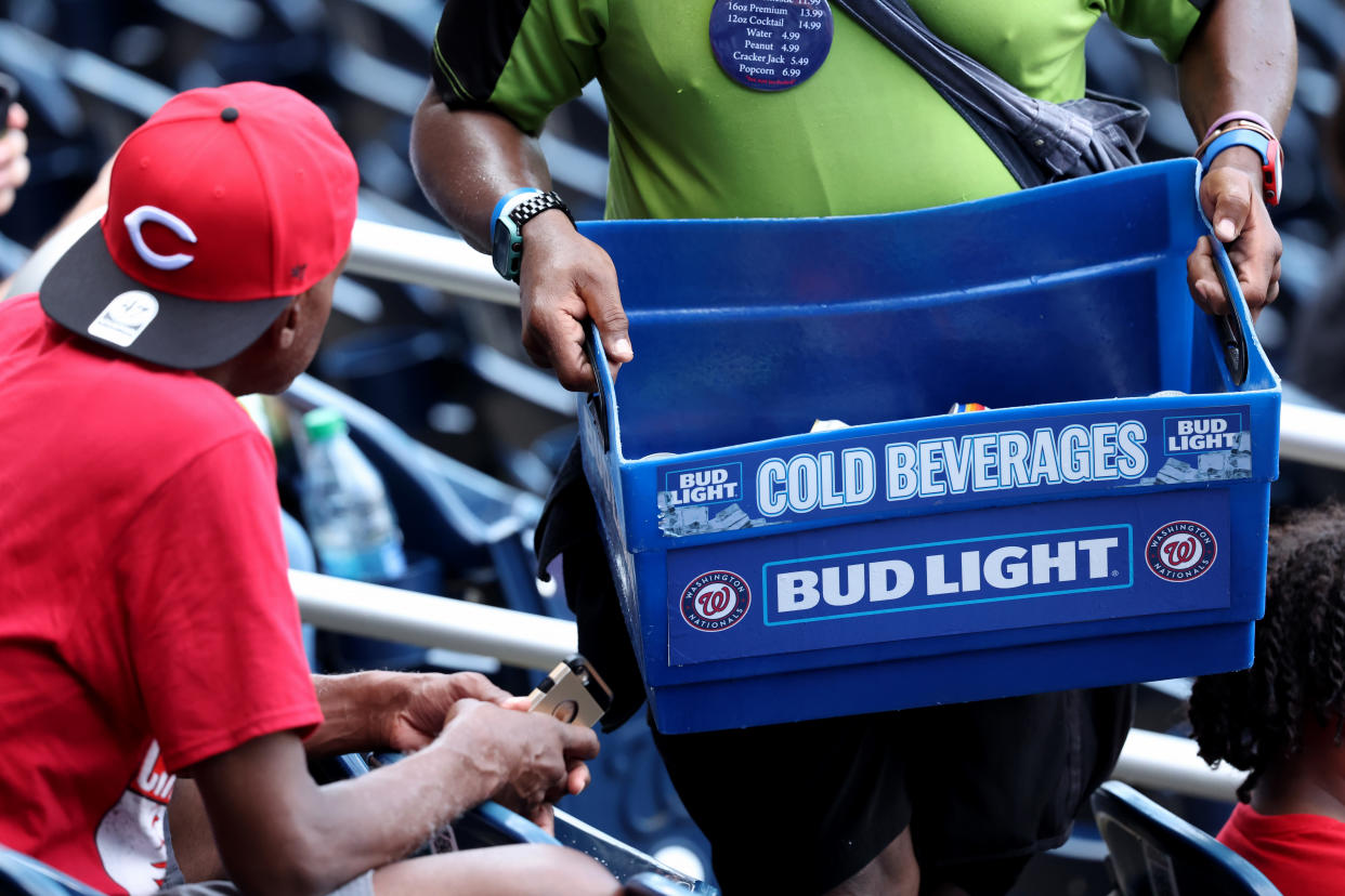 A beer vendor holds a mostly empty bin of Bud Light in front of a baseball fan wearing a Reds hat.