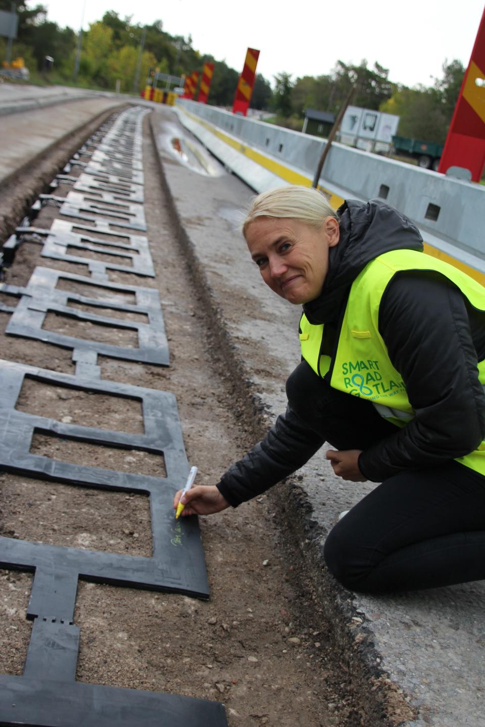 A woman in a yellow safety vest crouches beside road work, where startup Electreon is placing its black charging coils into the asphalt of a road in Sweden.