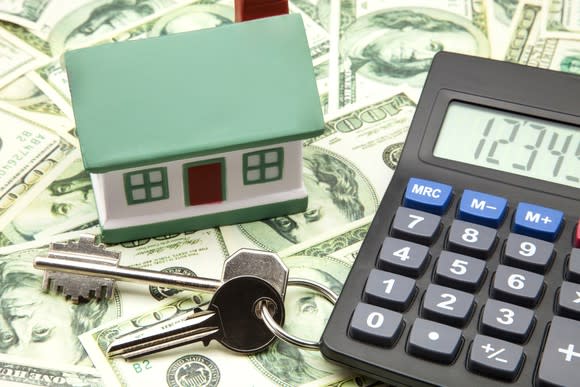 Toy house, calculator, and keys sitting on a flat surface on top of spread-out money.