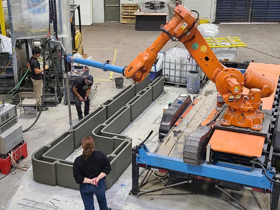 a 3D printer printing walls of a home in a factory near people