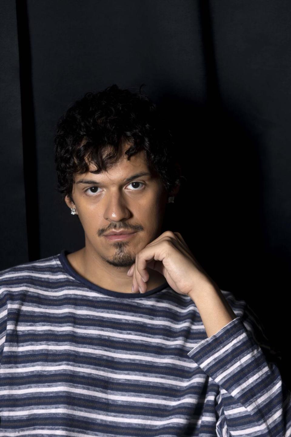 Omar Apollo in a striped shirt, facing the camera with his hand to his chin.