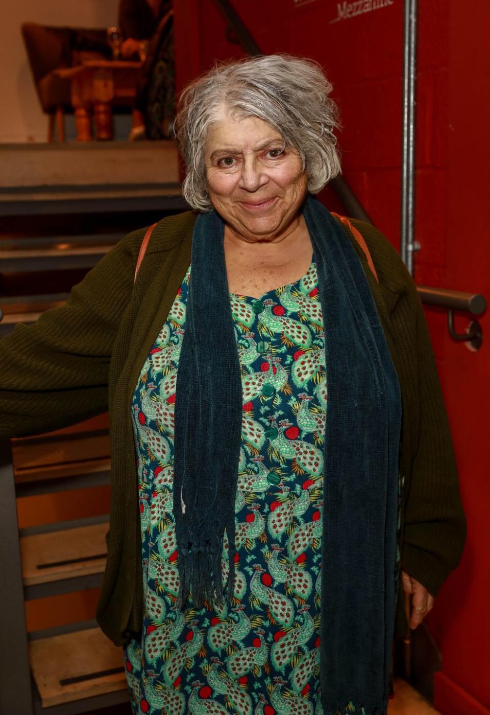 miriam margolyes in 2019, posing in front of a red wall, she is wearing a green cardigan and scarf and green and red dress and has short grey hair