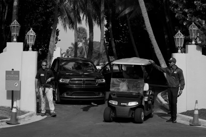 Two vehicles, a golf cart and a van, leave through the gates of Mar-a-Lago, which are flanked by two men in dark glasses.