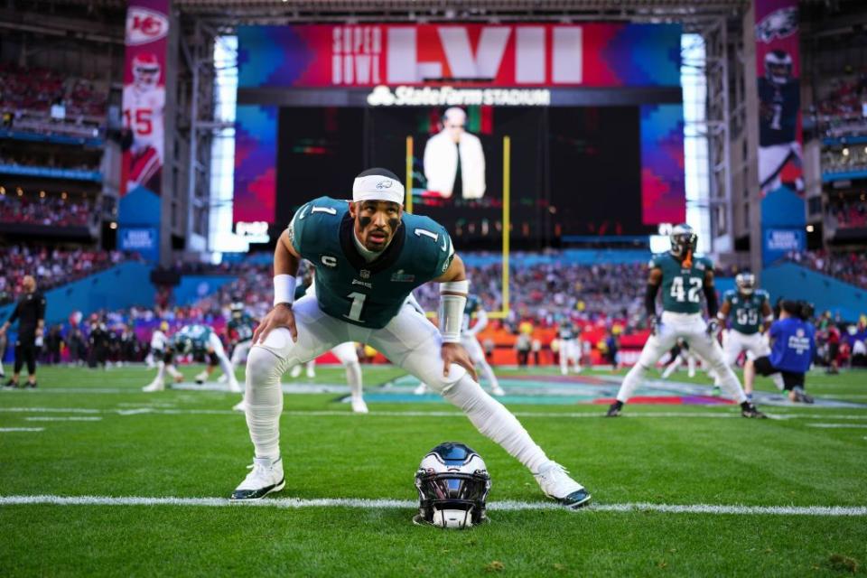 jalen hurts stands on a football field in a wide legged stretch, he wears a green and white football uniform with his helmet on the ground in front of him, other players are on the field in the distance, a large tv screen is also behind