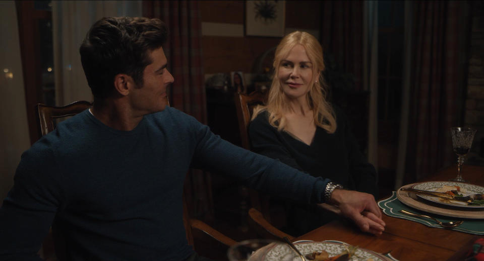 Zac Efron and Nicole Kidman are seated at a dining table, holding hands and smiling at each other. Plates of food are in front of them
