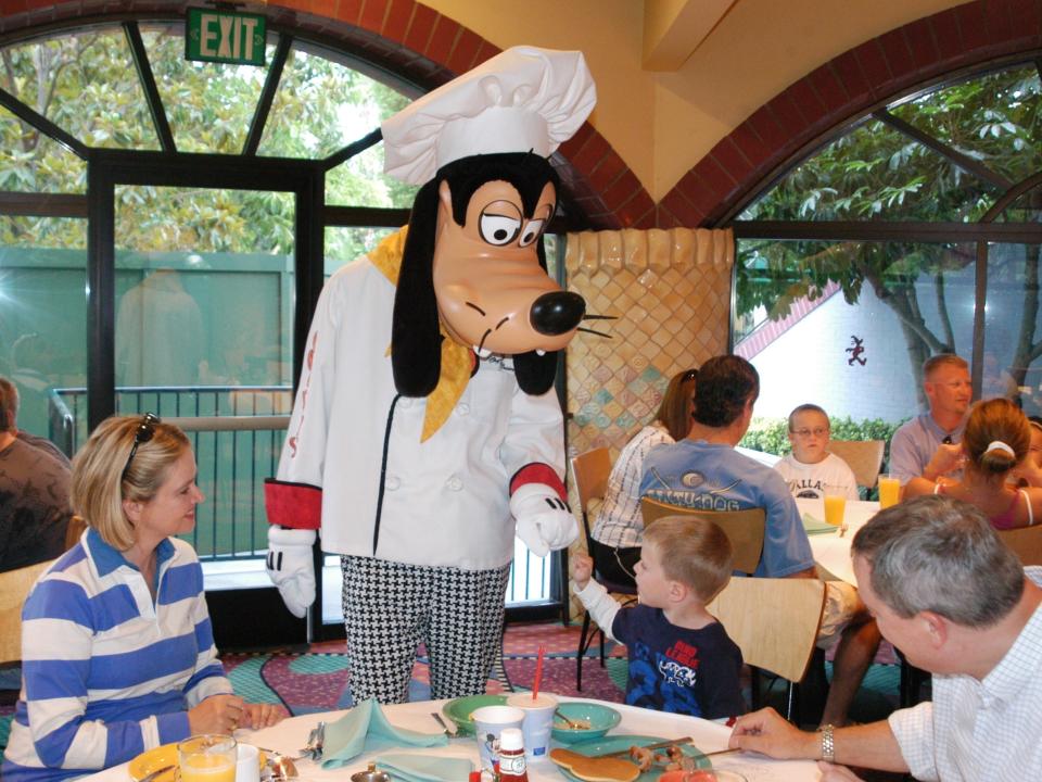 Someone in a Goofy dog costume in a chef's hat fist bumping a child sitting at a table in front of his breakfast with his family.