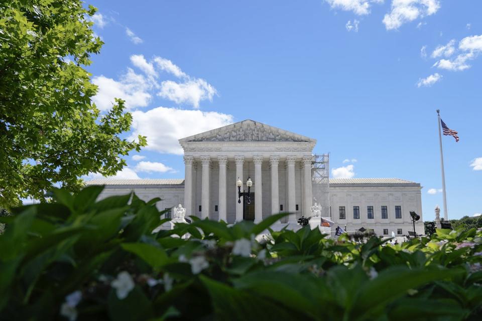 A white building with tall pillars, seen behind greenery and against a clear blue sky.