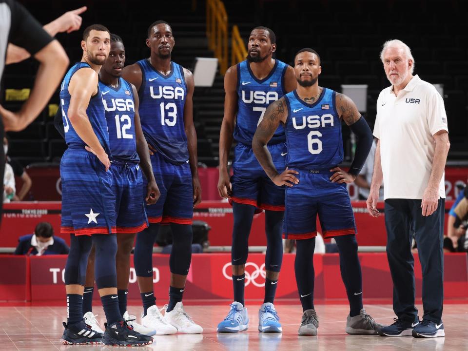 Zach LaVine, Jrue Holiday, Bam Adebayo, Kevin Durant, Damian Lillard, and Gregg Popovich stand during a basketball game at the Tokyo Olympics.