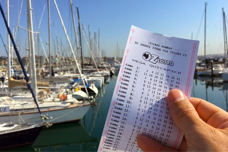 Hand holds OzLotto ticket in front of a marina. Source: The Lott