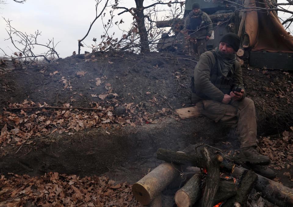 A Ukrainian soldier sits on a ridge in the ground in front of a small fire